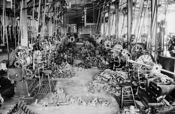 View of 21" and 24" lathes on the Willys-Overland Motor Company factory floor. These lathes machined rear axle parts, which are piled at each work station. The lathes are arranged end-to-end, and are attached to rotary belts which are suspended from the ceiling. Natural light is flooding the room from the factory windows.

The original envelope housing this photograph was inscribed with the following information: "Showing Gisholt Department of 9-21" Gisholt lathes and 1-24" Gisholt lathe. Since this picture was taken 4 more 21" Gisholt lathes have been installed in this department."