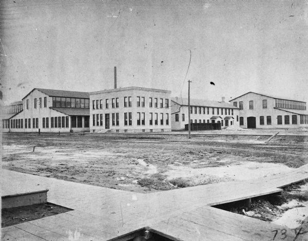 General view of the Gisholt Machine Company newly constructed main office (center) at 1315 East Washington Avenue, a locker room for workers (to the right of this office), machine shop (to the left with high pitched roof) and tool shop (to far right with high pitched roof). The wooden sidewalk in the foreground approximately marks the intersection of East Main and South Baldwin Streets in Madison. There is also a telephone pole and a smokestack.