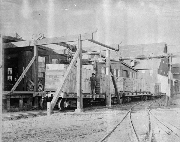 View of a loading platform by the railroad track at the first machine works plant of the Gisholt Machine Company, located at the 1400-block of East Mifflin and North Dickinson. Three flatcars are standing on the tracks adjacent to a warehouse loading dock, loaded with wooden crates of machines and manufactured parts on the decks. A worker is posing on one flatcar beside a massive crate, hoisted there by an overhead crane, with the stencil brand "From Gisholt Machine Co. Madison WI USA." The flatcars bear the initials, "C. & N-W. RY." for Chicago and North-Western Railway.