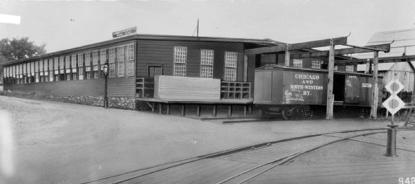 View across railroad tracks towards the loading platform at the first machine works plant of the Gisholt Machine Company located at the 1400 block of East Mifflin and North Dickinson. One railroad car for the "Chicago and North-Western Railway" is standing on the tracks adjacent to a warehouse loading dock, where there is a large crate. The car has chalk marks to indicate routing, contents, or other instructions. A sign on the top of the warehouse designates the manufacturing company and its signature product "Turret Lathes." The negative for this photograph has been retouched, particularly noticeable in the lamppost, window frames and railroad tracks.