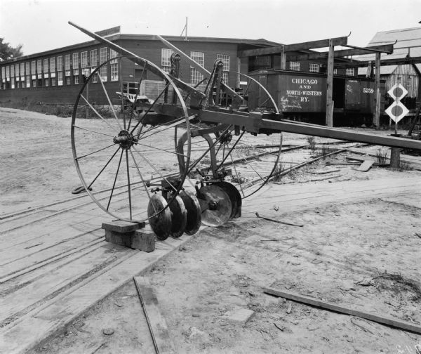 Horse-drawn harrow on display by the railroad yard platform at the first machine works plant of Gisholt Machine Company, located at the 1400-block of East Mifflin and North Dickinson. The agricultural tool is resting on the railroad ties in the foreground. In the background is the warehouse with a sign that reads: "Gisholt Machine Co. Turret Lathes." In front of the loading dock is a large crate and railroad car for the "Chicago and North-Western Railway." The car has been tagged with graffiti. 
