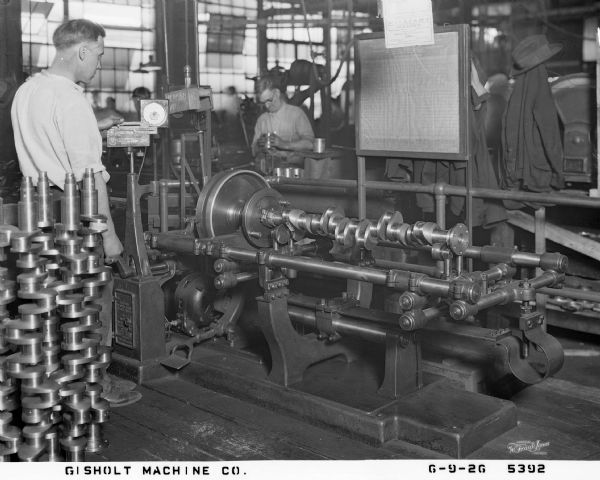 A worker is overseeing the production of crankshafts on Gisholt machinery at a plant for the Stutz Motor Car company. The Gisholt company logo is on the settings instrument and machine frame by each of his hands. Finished crankshafts are standing on end behind him. A worker across from him is standing at another work station looking down at a machined automotive part. Workers coats and hats are hanging on hooks and over posts nearby, and above the machined crankshaft is a sign for "Williams box lunch — Sandwiches, Fruit, Pie, Cake and Pickle" for 25 cents. 