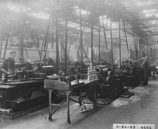Several machinists (some blurred by motion) are standing at work stations producing component parts on Gisholt lathes. This factory is one of countless companies that used Gisholt machinery for manufacturing in the United States and Europe.  
