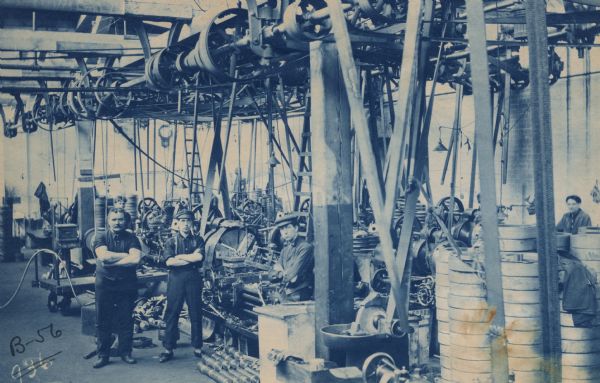 Factory workers standing with their arms crossed on the plant floor in the Gisholt Department of the Buick Company Plant #1 alongside lathes, and machined parts in piles and stacks. Radial belts and overhead electrical lights are suspended from the ceiling between work stations. The department floor shows ladders, a cart and clock. 

The Buick Motor Company was founded in Detroit, Michigan in 1903, and moved to Flint the next year. It began using Gisholt machinery at its automotive plants around 1910. 