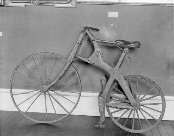 Wooden bicycle on display against a wall at the Wisconsin State Historical Museum, circa 1920. It is known as the "Crabtree Special" because it was reportedly made in 1891 from the crotch of a crabapple tree in Bunke Hill, Wisconsin, (near Platteville) by Walter Atkinson when he was 18 years old. According to descendants of Atkinson, Walter and his brother built the bike together. 

The bike was donated by Allan and Maurice Park to the Wisconsin State Historical Society circa 1916, when it became part of the museum collection. 