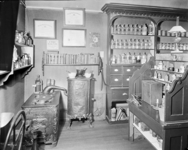 View of the pharmacy display at the Wisconsin Historical Society Museum. This installation with furnishings, equipment, and other stock supplies found in a druggist came from stores around Wisconsin. 

Three Certificates issued by the Wisconsin State Board of Pharmacy (one in 1911 for Charles LeComte in Green Bay) are hanging on the wall over a bookcase of publications on pharmacopeia. Several cabinets and wall shelves hold labelled glass jars of chemical ingredients used in making medicines. An incomplete pharmaceutical set up for distillation and steam (manufactured by F.A. Wolff and Sohne in Heilbronn) is standing in the corner; the distillation column is missing spouts on the two outlets at its base and a crankshaft for pulleys and a funnel at its top. Other objects used to produce medicines like mortars and pestles are also exhibited. An advertisement for "Dr. Twitchell's Excelsior Troches for Diptheria, Coughs, Hoarseness, Colds, Sore Throat, Catarrh, and Influenza" is tacked to the wall.