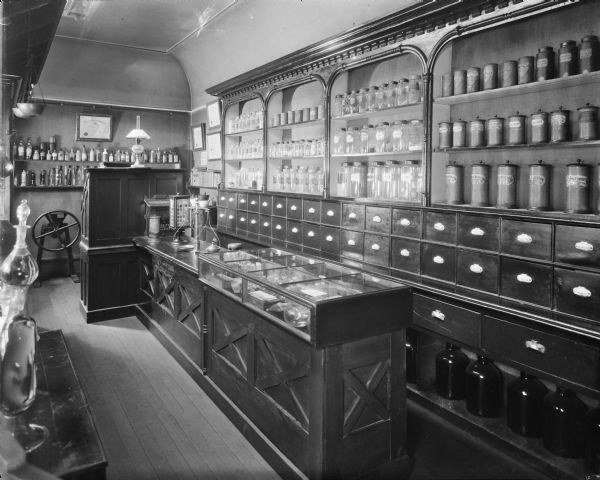 View of the pharmacy installation. It includes furnishings, equipment, and other stock supplies found at a druggist came from stores around Wisconsin.

Several cabinets and shelves hold labelled glass jars and lidded containers of chemical ingredients and prepared drugs. There are also countless drawers for filling prescriptions. In the middle of this exhibit is a long counter with glass display case to show other paraphernalia used by pharmacists, such as a brass scale with weights and measures, mortar and pestle, and fragile hand-blown glass instruments; there is also a rotating advertisement for Peerless Dyes. 