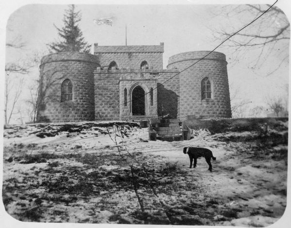 Winter view, with snow on the ground, of the Benjamin Walker Castle from the path leading up to the front entrance in the 900 block of East Gorham Street. There is a black dog on the path. 