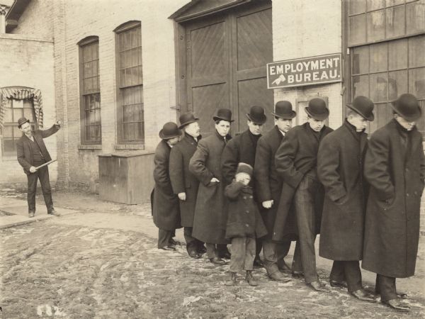The caption on the back of this photograph reads: "Eight of the Sales force of the Gisholt Machine Company. (Refused!)" It was made by "J.H.N." A line of eight men, who are wearing long overcoats and bowler hats, are walking in the plant yard in the opposite direction of a hand on a sign on the side of a building, "Employment Bureau." The line of men are walking with heads down, one holds the hand of a small child. They are walking away from a ninth man, who is in a suit and pointing at the group of men with a baton.