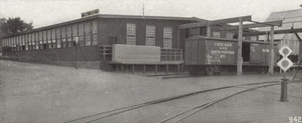 View across railroad tracks towards the loading dock at the first machine works plant of the Gisholt Machine Company, located at the 1400 block of East Mifflin and North Dickinson Streets. One railroad car for the "Chicago and North-Western Railway" is standing on the tracks adjacent to a warehouse loading dock where there is a large crate. The car has been tagged with graffiti. A sign on the top of the warehouse designates the manufacturing company and its signature product "Turret Lathes." 