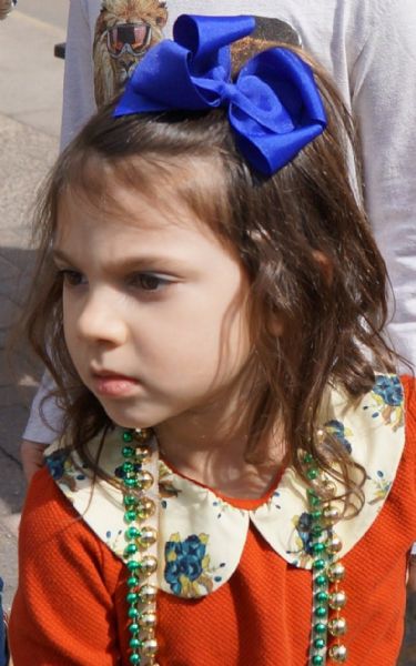 Quarter-length view of a young girl at the St. Patrick's Day parade. She is wearing a blue bow in her hair, a red dress with a white and blue floral print collar, and two strands of green and gold plastic beads.