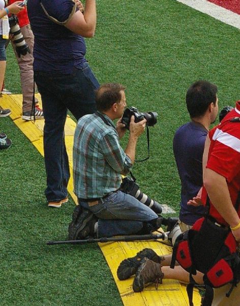 Slightly elevated view of Milwaukee Journal Sentinel photographer Richard (Rick) Wood. He is kneeling on the sidelines at a football game at Camp Randall Memorial Stadium,and is holding a camera in his hands. Another camera is in his lap, and a mono-pod is on the ground beside him. Other people with cameras are kneeling or standing around him. 