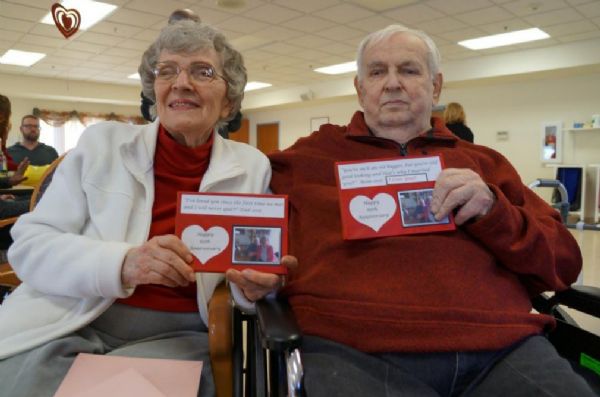 Portrait of David Joseph Marcou’s parents sitting side by side and holding up cards made by their daughter Lynn celebrating their 65th anniversary. 