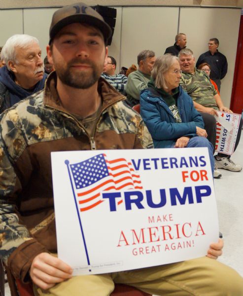 A young man is sitting in a chair and holding up a sign reading: "Veterans for Trump. Make America Great Again!" Other men and women are sitting in chairs behind him, and some of them are holding pro-Trump signs.