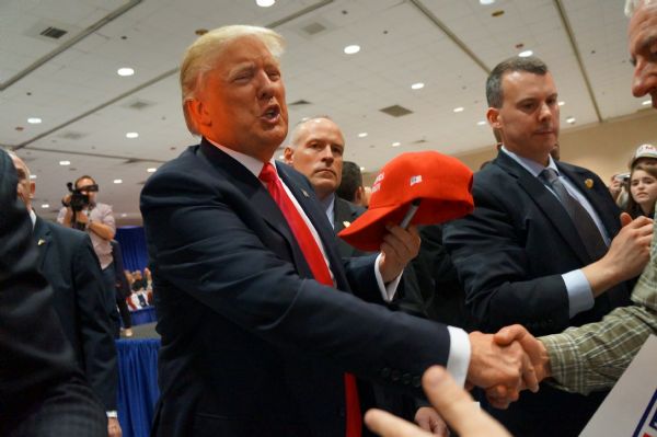 Donald Trump is holding a red "Make America Great Again" cap and shaking a man's hand.