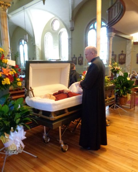 Father Robert Schaller is standing next to a casket holding the body of David Marcou's father, David A. Marcou inside St. James Church. There are flower arrangements placed on either side of the casket.