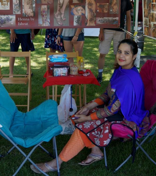 A young Indian woman, Ishrat, is sitting in a lawn chair in her booth at a fair. She is wearing a traditional Punjabi outfit (the salwar kameez) and is offering henna tattoos for sale at Riverfest held in Riverside Park. A large banner behind her showcases the various designs she has done in the past.