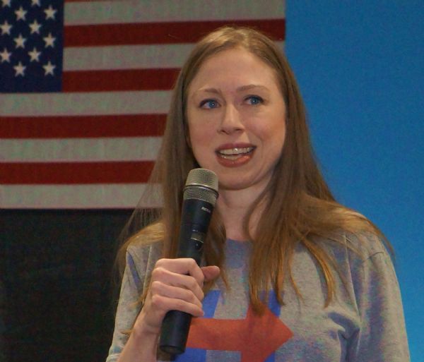 Head and shoulders view of Chelsea Clinton standing and speaking into a microphone at a Hillary Clinton rally at the University of Wisconsin-La Crosse. There is an American flag on the wall behind her and she is wearing a Hillary Clinton t-shirt.