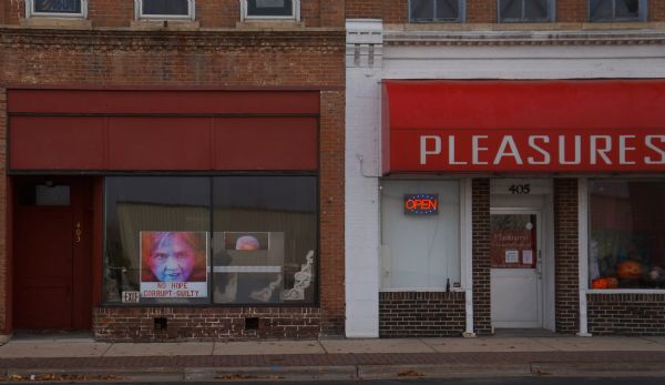 View from street towards two storefronts. The store on the left has a poster in the window with a red and blue image of Hillary Clinton and reads: "No Hope, Corrupt — Guilty." The store on the left is an adult store called: "Pleasures: A Lovers Boutique" and it is featuring Halloween decorations in the show window.