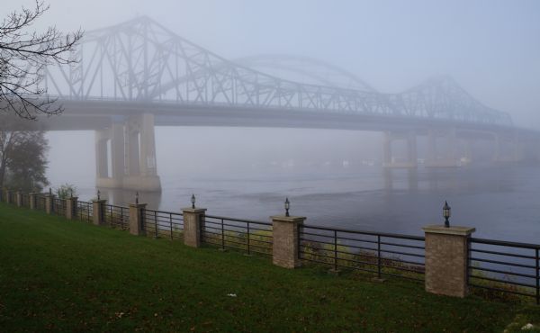 View across green grass and a fence looking towards the Mississippi River Bridge in the fog.