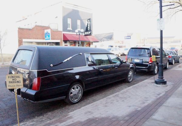 A hearse for David A. Marcou (David Joseph Marcou's father) parked along the curb outside of a sweet shop. Several signs are on the sidewalk that read: "Funeral, No Parking."