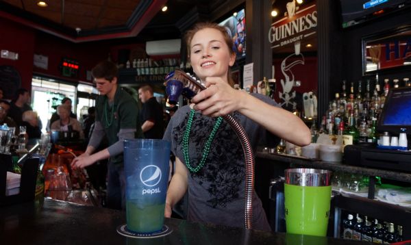 A young woman, Jamie, is pouring out a drink at Dublin Square Pub on the day of the St. Patrick's Day Parade. Both she and another bartender behind her are wearing green plastic bead necklaces. A crowd of people are sitting at the bar.