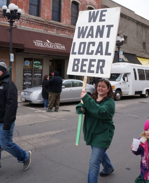 A woman in a green sweatshirt is marching in the St. Patrick's Day parade carrying a sign reading: "We Want Local Beer."