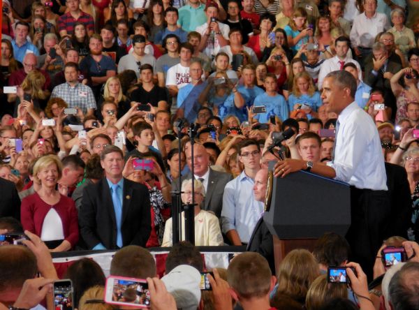 President Barack Obama is standing at a podium delivering a speech at UW-LaCrosse. The crowd is filled with mostly young men and women, many of whom are holding up their cellphones to take pictures or videos. In the front row on the left are Senator Tammy Baldwin and Congressman Ron Kind.