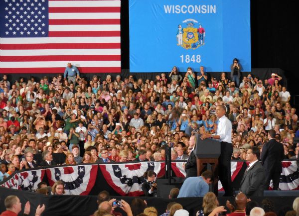 President Barack Obama is standing at a podium and addressing a crowd of people at UW-LaCrosse. Three secret service men are standing around him. Both the American flag and Wisconsin flag are hanging on the wall behind the crowd. Senator Tammy Baldwin and Congressman Ron Kind are sitting in the front row in the center.