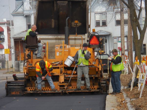Four men are working on the back of a paver as it is laying asphalt on 7th Street. The men are all wearing reflective vests. 