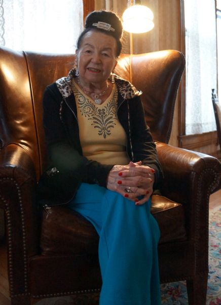 Indoor portrait of Magda Herzberger, a holocaust survivor and author, at Wheeler House. She is sitting in a leather chair, and is wearing a yellow shirt, blue pants, and a black hoodie with leopard print lining. She is also wearing a heart-shaped necklace, earrings, and a decorative headband to hold up her hair in a bun. 