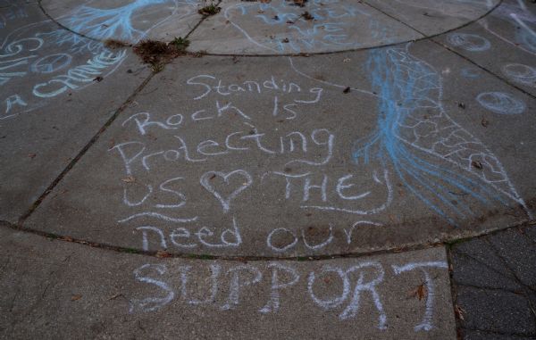 View of a message written in chalk on a sidewalk at Riverside Park. It reads: "Standing Rock is Protecting Us [image of a heart] They need our support." There are other drawings and messages in chalk nearby.