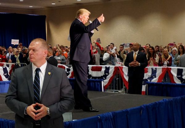 Donald Trump is standing on a stage holding a microphone in one hand and giving a thumbs up with the other hand. Two secret service men are standing on either side of the stage. The audience is behind a barrier with red, white and blue bunting.