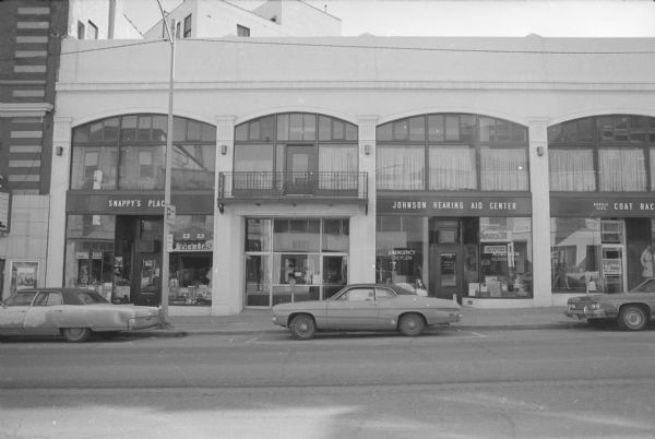 View across King Street towards the Arcade building. The right side of the Majestic theater building is on the far left, and then the storefronts are, from left to right: Snappy's Place, Norris Lea Furs, Johnson Hearing Aid Center, and Lea's Coat Rack. Storefronts on the other side of the street are reflected in the show windows, including The Gallery Inn restaurant and Econo Print.