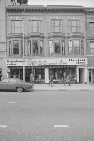 View across North Carroll Street towards the Spoo and Son storefront, with signs for Hickey-Freeman clothes, and Florsheim shoes. There are mannequins in the windows wearing suits and ties. Next door on the left is the Gay Building, and next door on the right is the Karstens storefront.