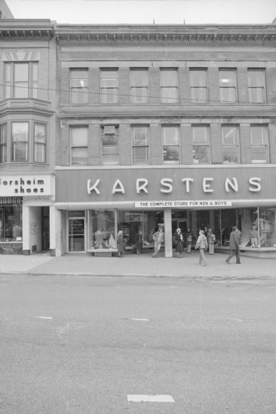View across North Carroll Street towards the Vroman block. A glass door on the far left has a sign for the Credit Bureau of Madison. A large sign above the show windows reads: "Karstens, the Complete Store for Men & Boys." Pedestrians are standing under the recessed entryway near the show windows and a column. Next door on the left is the Spoo and Son storefront.