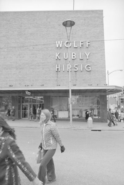 View across intersection of North Carroll Street and State Street towards the Wolff, Kubly and Hirsig Company building. Pedestrians are in the foreground, and on the sidewalk. On display in the show windows are Speed Queen washers and dryers.