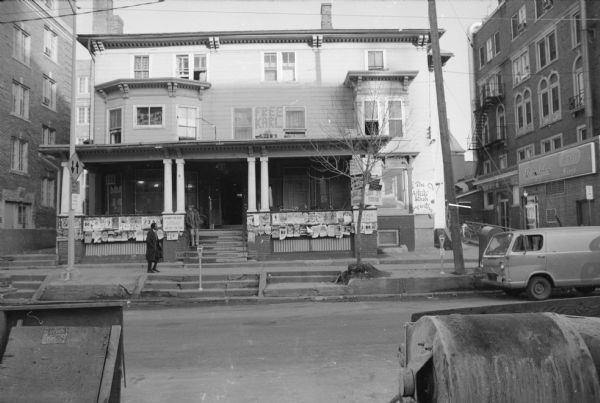 View across Lake Street towards the Lake Street Station. Signs and posters line the porch, including one advertising rooms for rent. One sign on the far right reads: "The Artists' Stash Cooperative." Graffiti is above the porch on the siding between two windows, and reads: "Free Karl." A person is standing on the front steps leaning against the columns. Another man is walking on the sidewalk in front of the house. The building on the right across the alley has a sign on the side for the Rennebohm Drug Store.