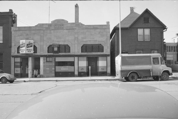 View across North Henry Street towards the rear of the Belle Osborn building (left) and a wood frame house (right). The house is the backside of Parthenon Gyros. The Belle Osborn building contains DiSalvo's Villa, a tavern, with windows and signs advertising that they sell Schlitz beer. A van is parked outside of the wood frame house. 
