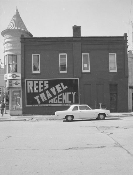 View across North Henry Street towards the Matthew Gay Building, which has a turret above the corner entrance, at the intersection of State Street. On the ground floor is the Triangle Superette. Signs above the show windows read: "Pepsi" and "Pepsi-Cola." The billboard on the side of the building reads: "Rees Travel Agency, 330 State St." A car is parked along the curb next to a meter.