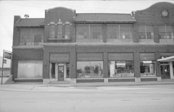 View across West Johnson Street towards Madison Motors. Signs in the windows, and on a post near the left side of the building, are advertising Dodge Parts and Services. In the show windows are parts and accessories. The windows are reflecting the buildings that are across the street.