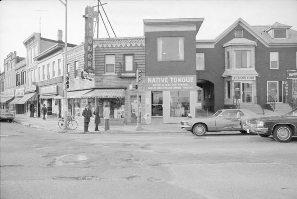 View across West Gorham Street at the corner of State Street, towards Badger Liquor and Native Tongue Indian & Mexican Apparel. A large neon sign is at the corner of Badger Liquor. Pedestrians and cars are waiting for the traffic lights to change. To the right of Native Tongue is a Veterinary Clinic. To the left of Badger Liquor is a view down State Street.