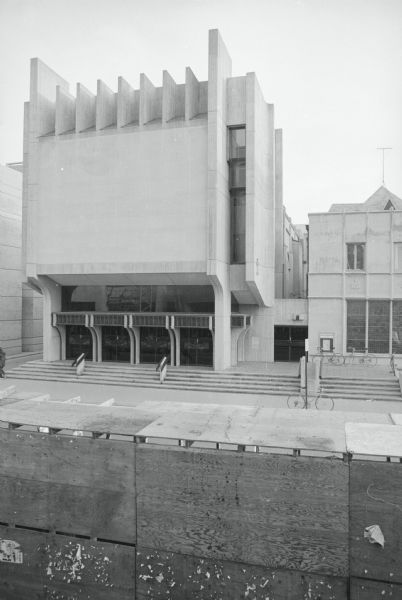 Slightly elevated view across State Street towards St. Paul's University Catholic Church, a Brutalist style of architecture. Several bicycles are parked outdoors to the right of the church. A wooden barricade for the expansion of the Memorial Library is in the foreground.