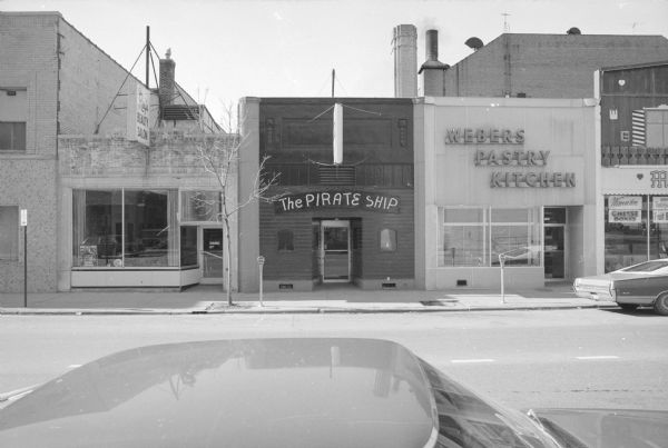 View across North Fairchild Street towards a row of businesses. From left to right: Regis Beauty Salon, The Pirate Ship (a bar), and Webers Pastry Kitchen. Miller's International Market is to the right of Webers, and the Madison Community Center is to the left of the Regis Beauty Salon. An automobile is parked in the foreground.