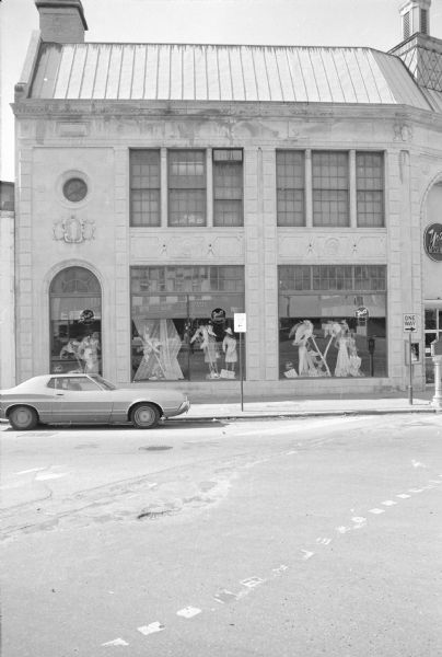 View across North Fairchild Street towards Yost-Kessinich Department Store on the corner of State Street. Show windows display mannequins in women's clothing among fake pine trees. The windows are reflecting the businesses across the street. To the left of the building is Miller's International Market.