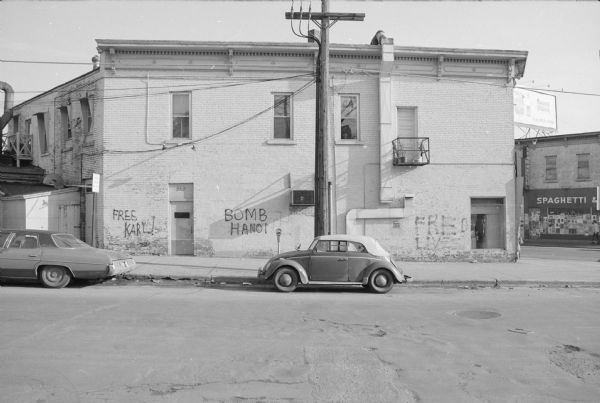 View across West Gilman Street towards the Chocolate House on the corner of State Street. The side door on the left leads up to apartments on the second floor. Graffiti is sprayed on the wall, reading, from left to right: "Free Karl!" "Bomb Hanoi" [with faded swastika symbol next to it], "Fred Lives." The storefront on State Street on the far right has a sign that reads, in part: "Spaghetti &," and a billboard above is for "Arandas Tequila."