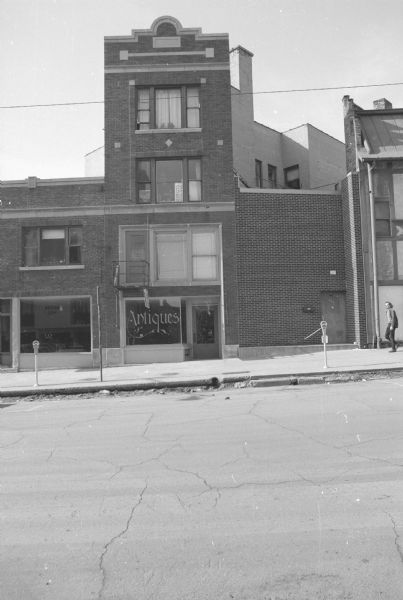 View across West Gilman Street towards an antique store. The upper floors hold apartments. A reflection of the Master Hall across the street is reflected in the ground floor window on the left. On the third floor of the buildings is a sign, sideways in the window, that reads: "Service Window Closed." A man on the far right is walking along the sidewalk.