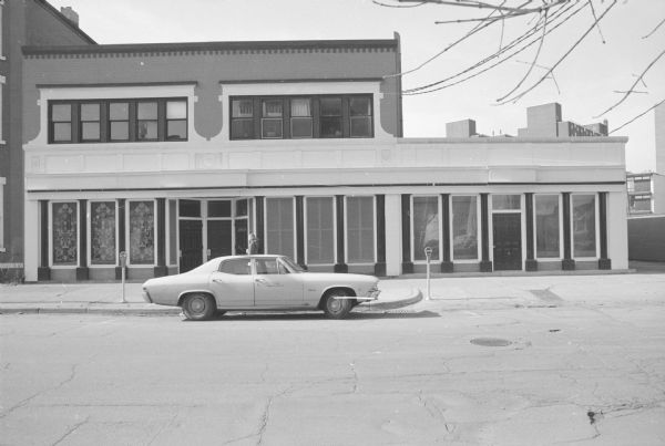View across West Gilman Street towards the George E. Leonard Store Building. The offices or stores inside the building are unknown. The three ground floor windows to the left of the doors are decorated with an ornate floral pattern. A woman is walking on the sidewalk near the recessed entrance to the building, just behind a car parked along the curb. 