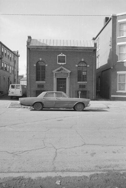 View across West Gilman Street towards the Grimm Book Bindery. Two plaques are on either side of the door, and a sign above the doorway, all read: "Grimm Book Bindery." A car is parked along the curb in front of the building, and a van is parked in an alley on the left. 