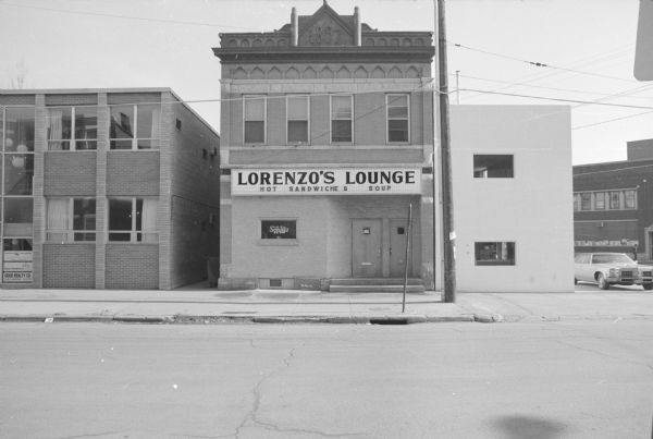 View across West Gilman Street, towards a brick building near the corner of University Avenue. A marquee above the door reads: "Lorenzo's Lounge: Hot Sandwiches, Soup." A neon sign in the window advertises Schlitz beer. Above the second story windows is decorative brickwork, and a date stone reads: "1897" at the roofline.